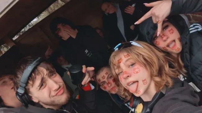 A group of people inside a wooden shelter, posing for a selfie. Several individuals have stickers of red cartoon cat faces on their faces. The person in the foreground on the left wears a headset and makes a pouty face. The person on the right sticks out their tongue and makes a peace sign. Other people in the background are making various expressions and poses.