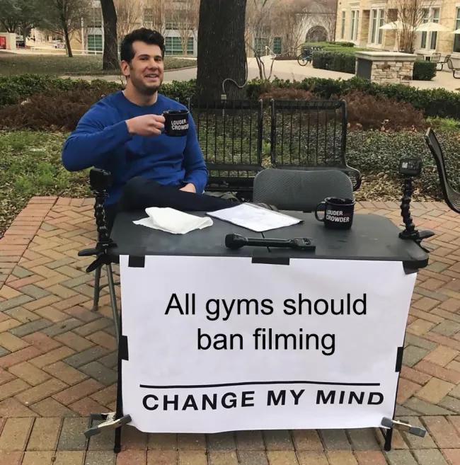 A picture of a man sitting behind a table outdoors smiling. Tapped to the front of the table is a white banner which says 'All gyms should ban filming. Change my mind'