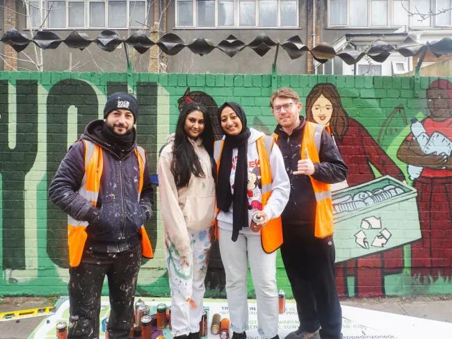 A group of young people, two men and two women, stand in front of brick wall that has been painted with street art to depict recycling empty containers. There are lots of spray cans on the floor around them.