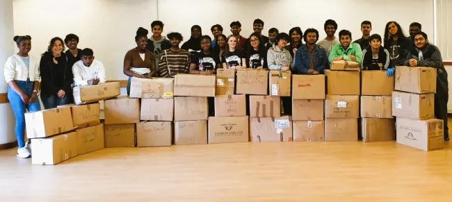 A group of young people standing behind stacked boxes full of clothes and shoes that had been donated to send to Ukraine.