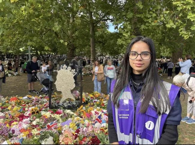 Shamza in a purple vest standing near a floral tribute to Queen Elizabeth II in a park filled with flowers surrounded by people.
