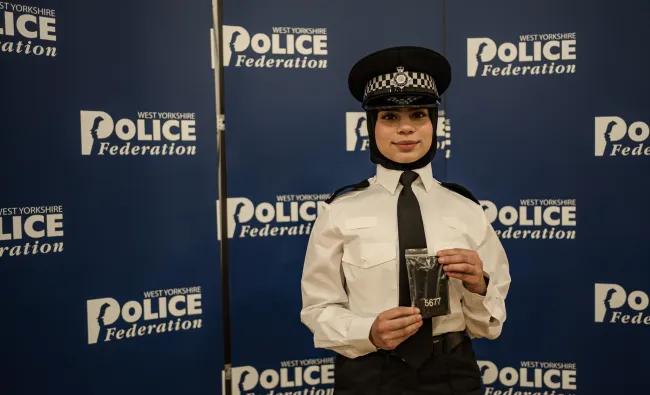 Shamza, a female police officer in uniform holding a service badge in front of a West Yorkshire Police Federation backdrop.