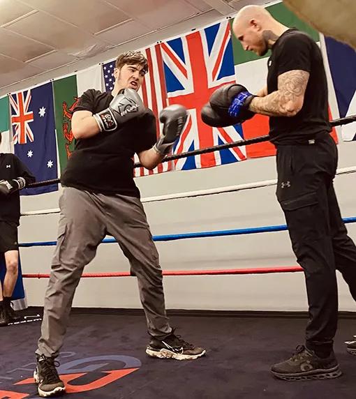Simon is practising boxing with his coach. They're both standing in a boxing ring, Simon has boxing gloves on in a boxing stance, using his hands and arms to protect his body. His coach, Luke, is wearing boxing pads for Simon to practice throwing punches on.