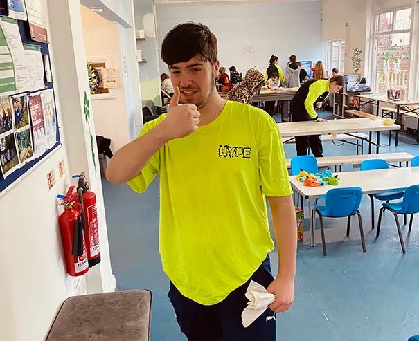 Simon wearing a bright yellow shirt with the word "HYPE" on the shirt, giving a thumbs-up while standing in a youth club hub centre.
