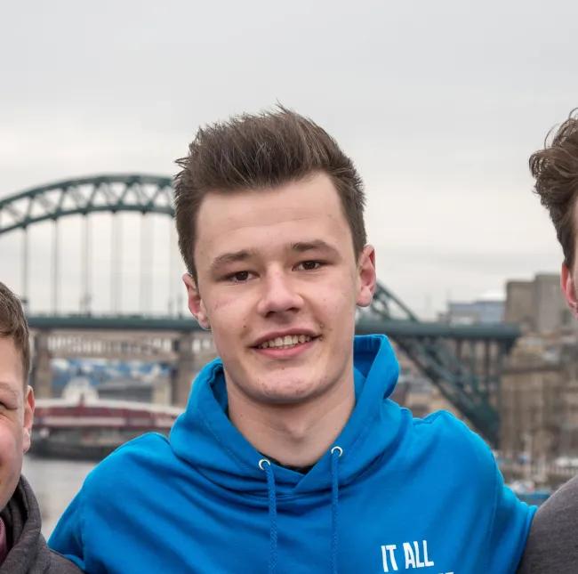 A close-up picture of Kai outdoors, wearing a bright blue hoodie with white text that reads "It All Starts at Yes." He is smiling with a bridge and city buildings blurred in the background.