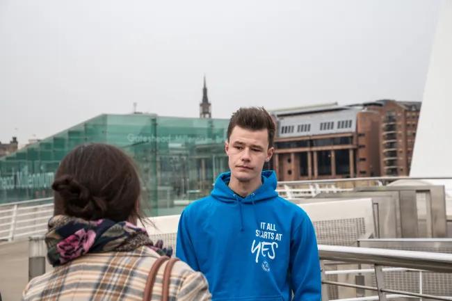 Kai outdoors wearing a bright blue hoodie with white text that reads "It All Starts at Yes." He is having a conversation with a woman with her back to the camera, wearing a brown stripped shirt with a scarf covering her neck. 