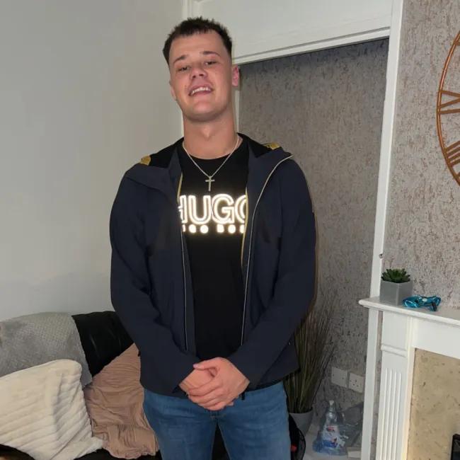  Kai dressed casually in a dark jacket over a black Hugo Boss t-shirt, standing in a living room. He is smiling with his hands clasped in front of him. 