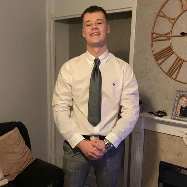 Kai dressed in a white dress shirt with a black tie and grey checked trousers, standing confidently in a living room. He is smiling, with his hands clasped in front of him. 