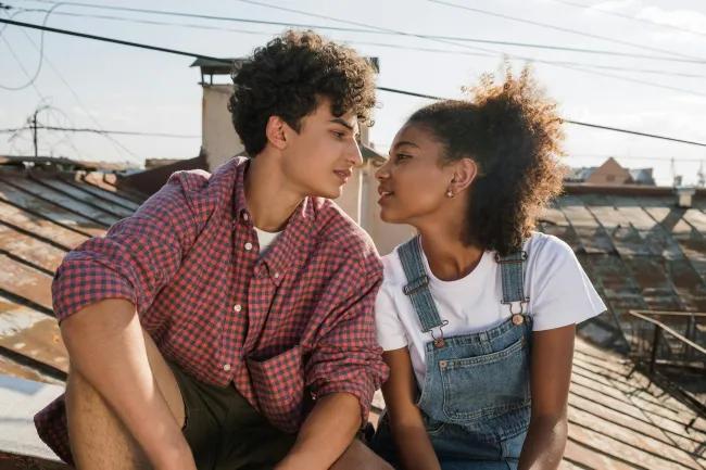 Two teenagers sitting on a roof looking into each others eyes and leaning towards each other.